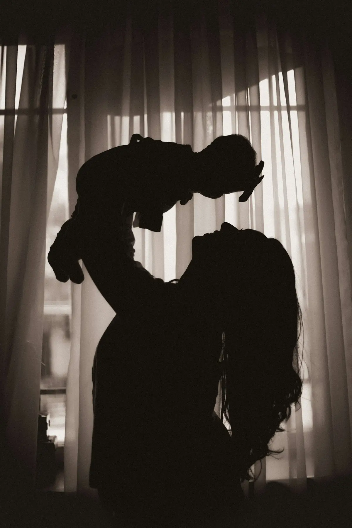 Silhouette d'une mère soulevant son bébé devant une fenêtre.